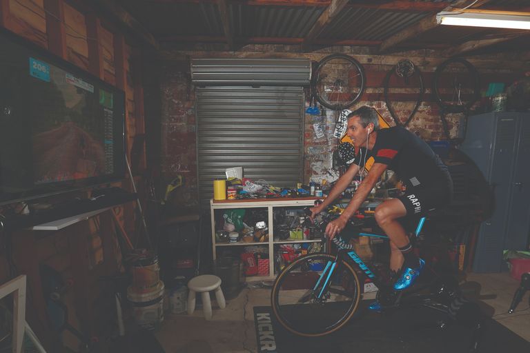 Best cycling headphones allow for total immersion in the ride. This image shows a rider to the right of the image, on his bike on a turbo with headphones on, looking at a big screen in a garage. In the background is a shelving unit and metal roller shutter with wheels hanging up. 