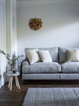 grey sofa in gray and white painted living room, gray graphic rug, dark hardwood floor, rattan pendant light, small stool with vase and tub, cream cushions