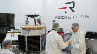 white coat lab workers carry a pain of glass near a golf-cart-sized satellite in a Rocket Lab branded white room.