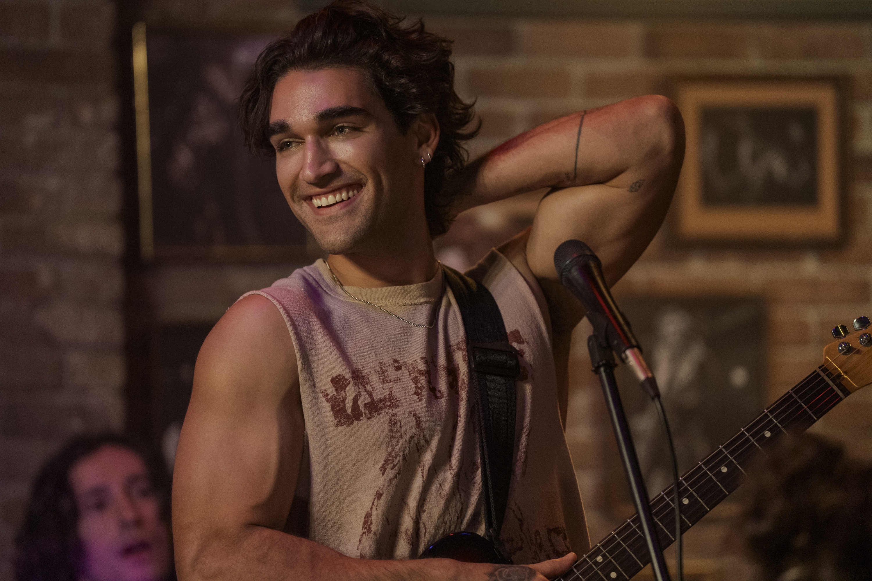 Justin (Josh Heuston) smiles and lifts his arm overhead while on stage playing guitar at a venue in off campus season 1