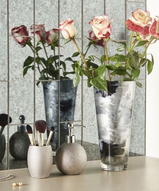 A greige bathroom shelf with a silver pot of makeup brushes, a silver soap dispenser, and a silver vase of pink roses, with mirrored wall paneling at the back