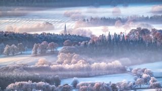 Frosty and misty landscape of the Peak District, with a church spire in the far distance surrounded by layers of open fields and forest