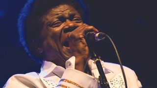 Charles Bradley on stage in a white sequinned jacket, singing into a microphone.