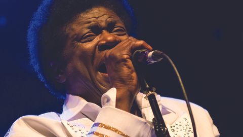 Charles Bradley on stage in a white sequinned jacket, singing into a microphone.