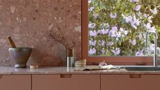 Close-up image of a kitchen sink and countertop. There is a window behind the sink and the countertop is made out of blush pink terrazzo. The cabinetry is made of a matching blush pink color. 