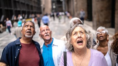 A group of older people stand in the middle of a city street, looking up in fright. It looks like a still from a horror film.