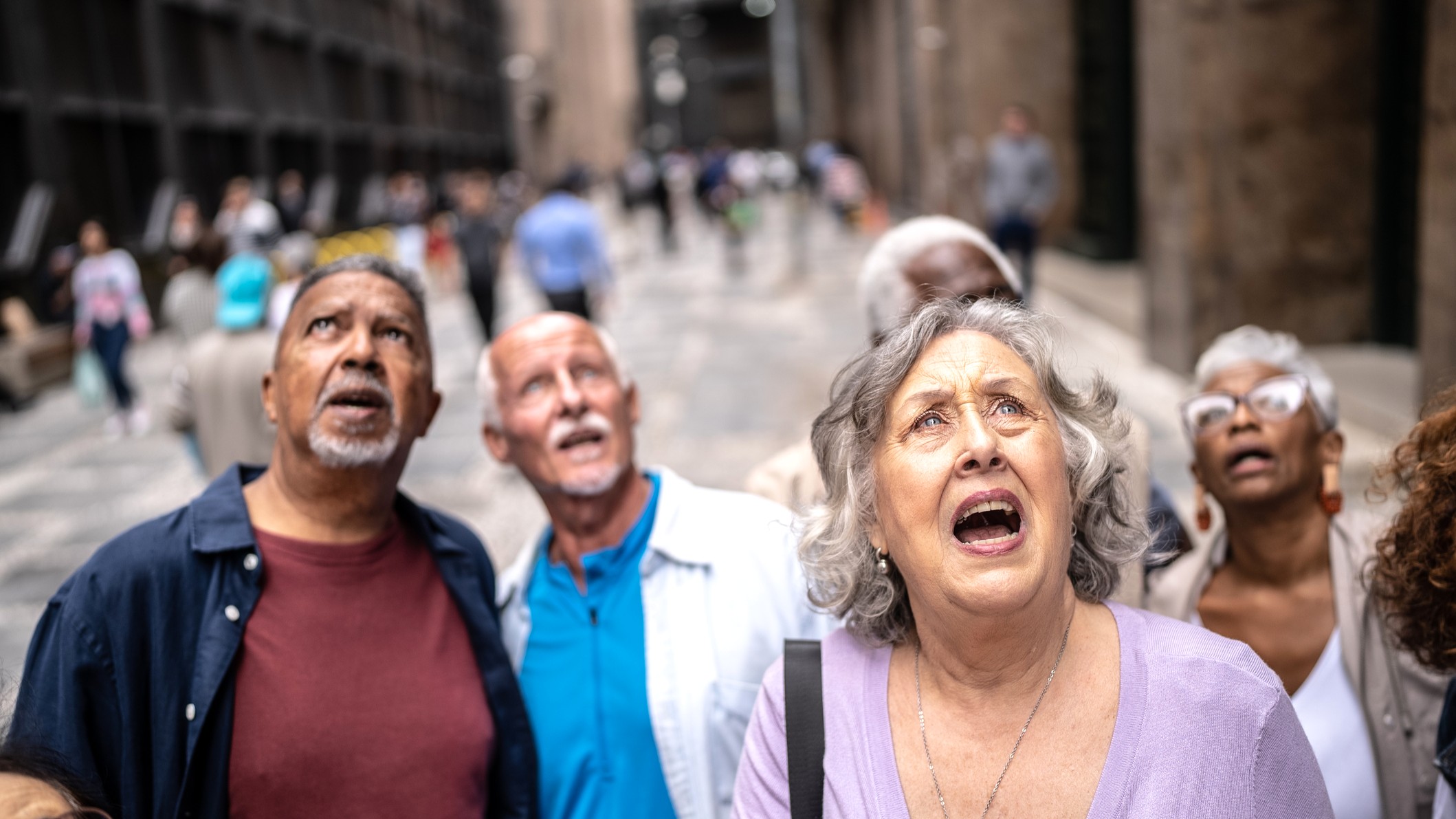 A group of older people stand in the middle of a city street, looking up in fright. It looks like a still from a horror film.