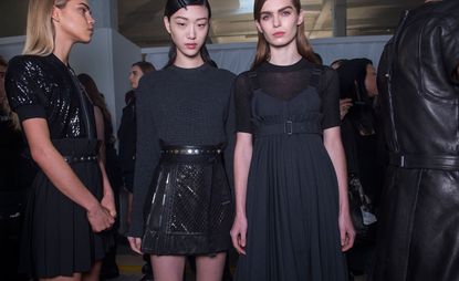 Three female models wearing looks from the Diesel Black Gold collection. One model is wearing a black sequin top, short black pleated skirt and embellished belt. Next to her is a model wearing a dark grey long sleeve sweater and a short black embellished skirt. The third model is wearing a semi-transparent top and grey pleated dress. And there is a fourth person at the edge of the photo wearing a black leather piece