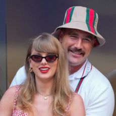 Taylor Swift wears sunglasses and bright red lipstick while Travis Kelce puts his arms around her while wearing a Gucci bucket hat