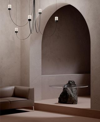 A render showing Paul Cocksedge's Gravity Chandelier for Moooi, a design featuring black curved arms hanging from the ceiling in a taupe-coloured room with an arched door, a side table made of stone and glass and a leather sofa