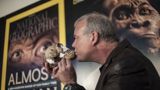 A man in a suit kisses the mouth of a Homo naledi skull. Behind him are posters featuring Homo naledi.