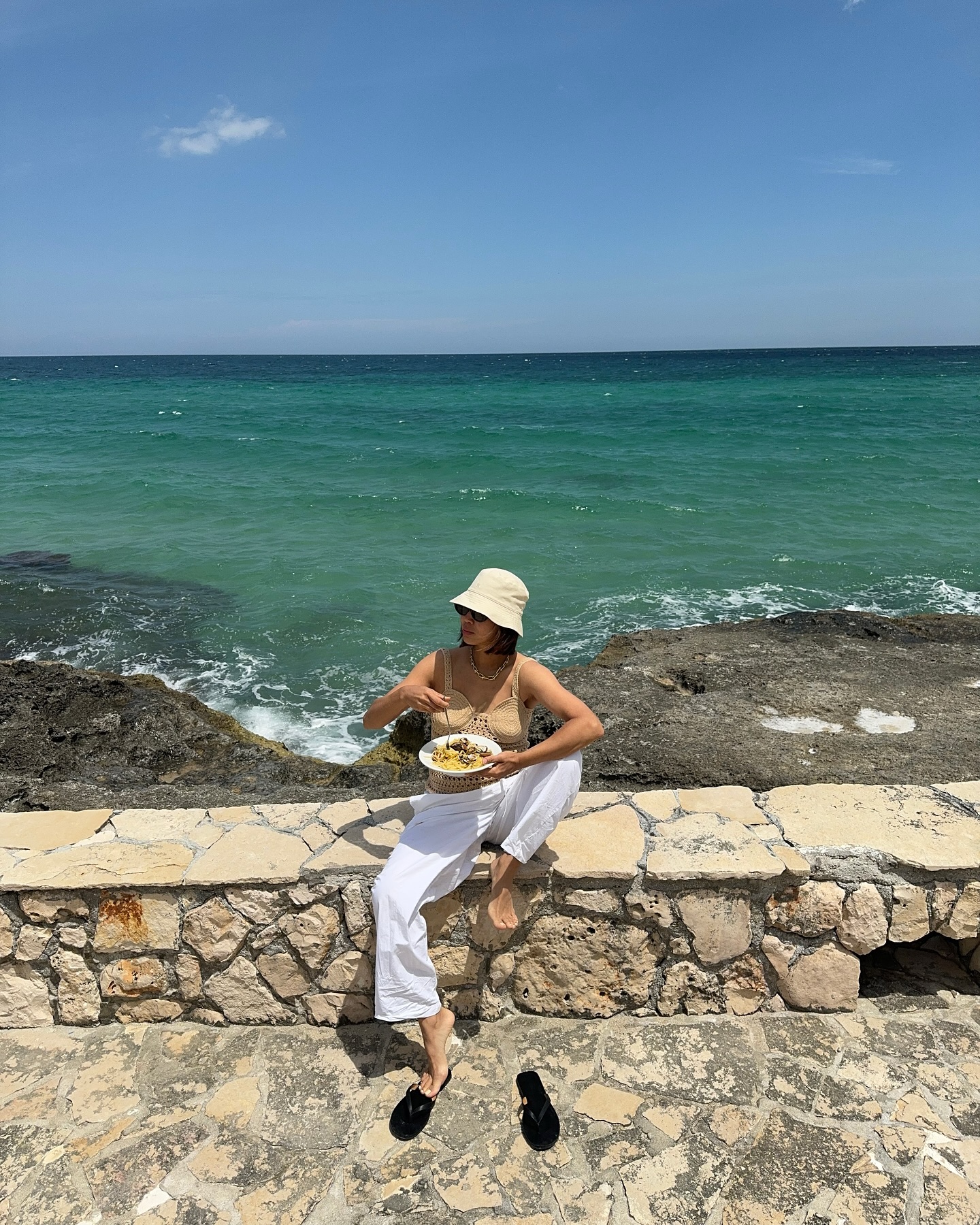 An image of @aimeesong wearing a holiday outfit consisting of a bucket hat, white linen pants, and beige tank top.