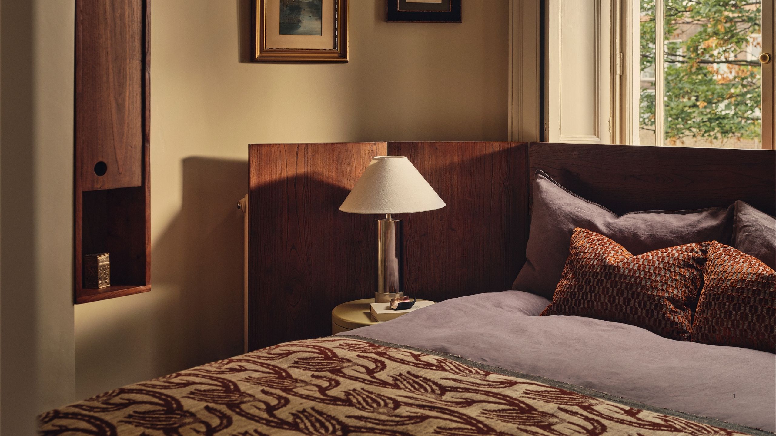 Image of an off-white bedroom with a large bed that has an extended wooden headboard. The bedding is purple and red, and there is a lamp by the bed.