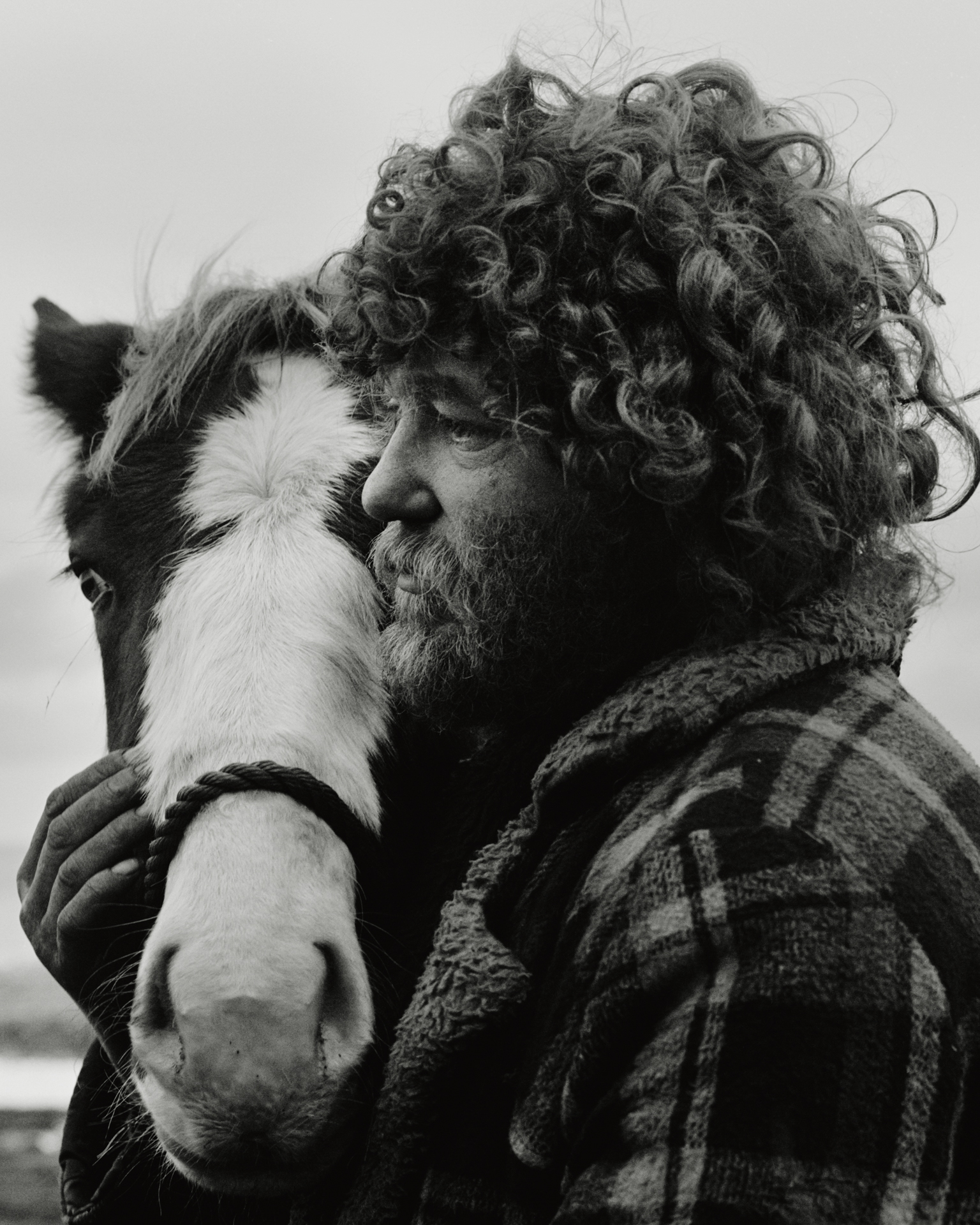 Black and white portrait of a man with curly hair and a full beard pressing his face close to a piebald pony's head, taken on Seacoal beach, Lynemouth, Northumberland, in 1983.