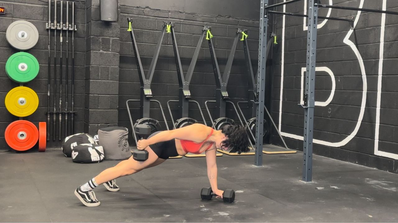 Raquel Sanjurjo performs a renegade row into triceps kickback in a gym. She is in a high plank position holding a pair of dumbbells. One is on the floor, the other is held next to her thigh, with a straight arm.