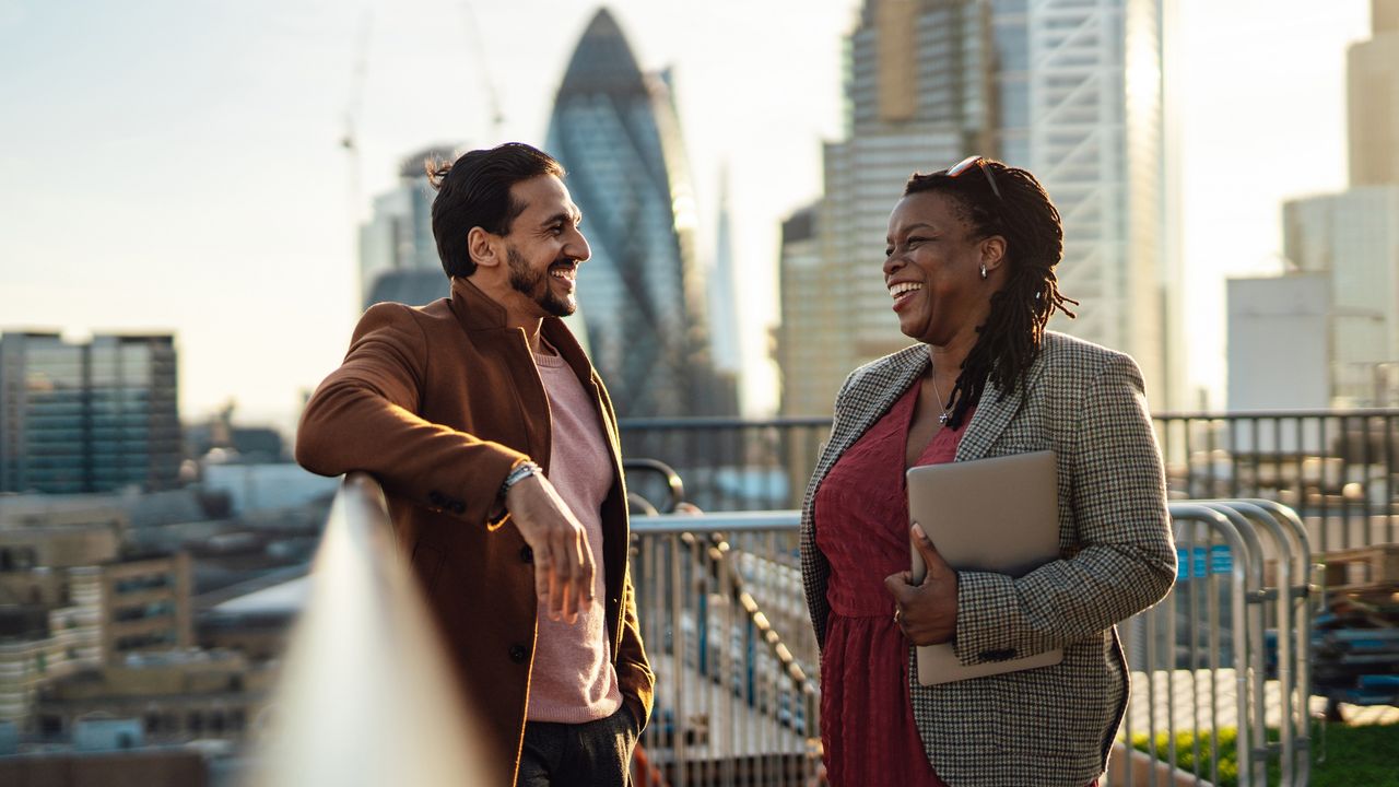 Two executives smile while chatting on the roof of a tall building with a city skyline in the background.