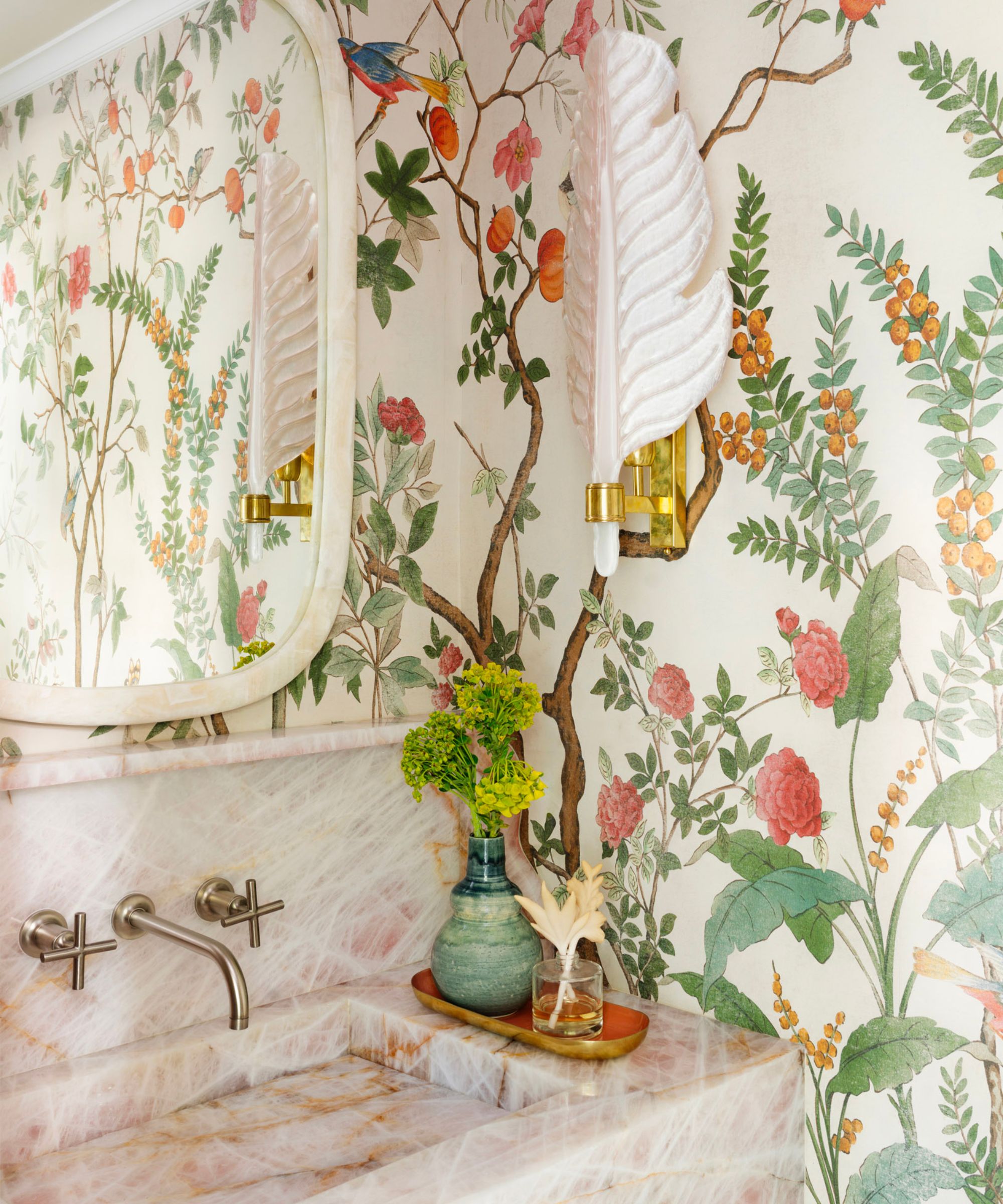 The corner of a bathroom with a pink marble sink, floral wallpaper, a soft pink leaf wall sconce, and a mirror.