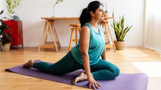 Woman at home in pigeon pose. She is on a purple yoga mat with one leg extended behind her and the other in front of her, with her knee bent and her left foot under right hip.