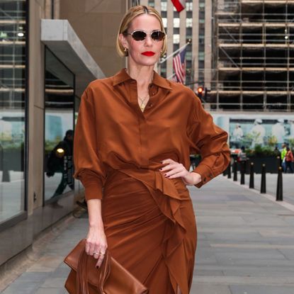 Leslie Bibb wearing a bob hairstyle and a monochromatic tan outfit. She is wearing oval sunglasses and a red lip. 