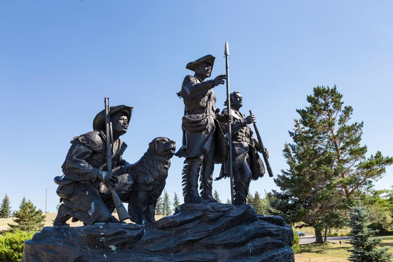 A bronze statue depicting three explorers and a dog standing together on a rocky plinth beneath a clear sky.
