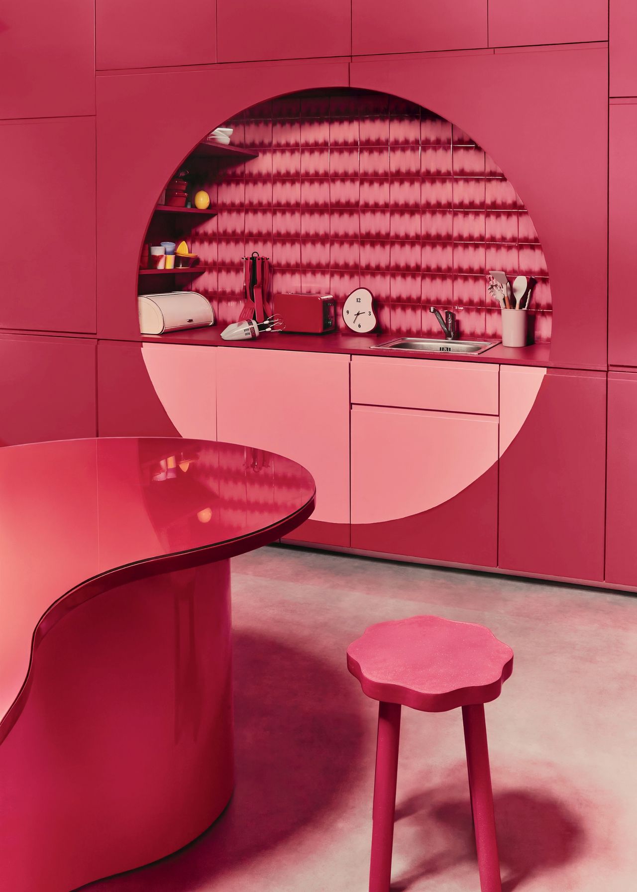 a two-tone pink kitchen space with a large circle design over the counter hiding storage behind either edge for shelving with a circular, wavy table with a stool in front on concrete flooring