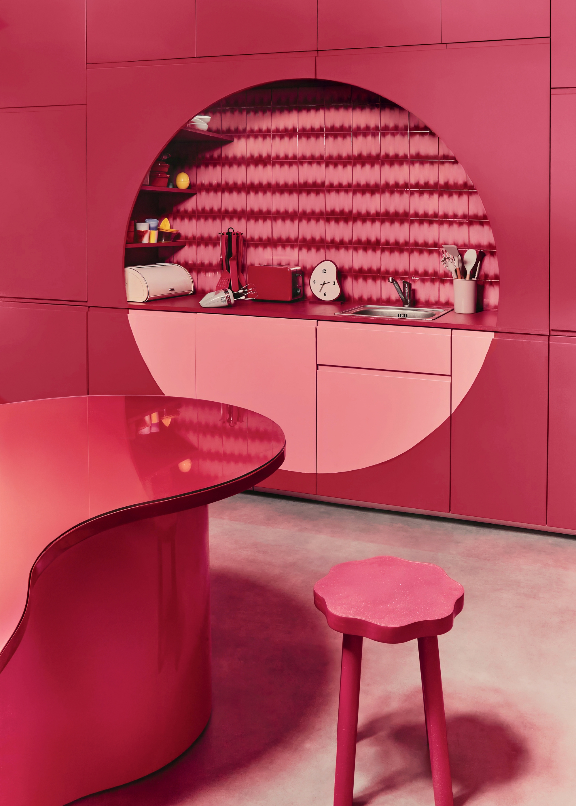 a two-tone pink kitchen space with a large circle design over the counter hiding storage behind either edge for shelving with a circular, wavy table with a stool in front on concrete flooring