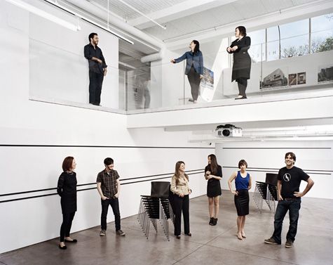 An open area in a room with 2 floors , white walls and ceiling. A man and 2 women standing on the top balcony (made of clear glass). Below them on the bottom floor is filled with 6 people standing in the room withare 2 sets of stacked black chairs with metal legs, and a projector overhead