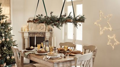 a festive dining room with beige walls, a wooden and beige dining set decorating with lots of Christmas ornaments, a fireplace filled with festive candles, a garland hanging above the table, a Christmas tree in the corner, and two star lights on the wall