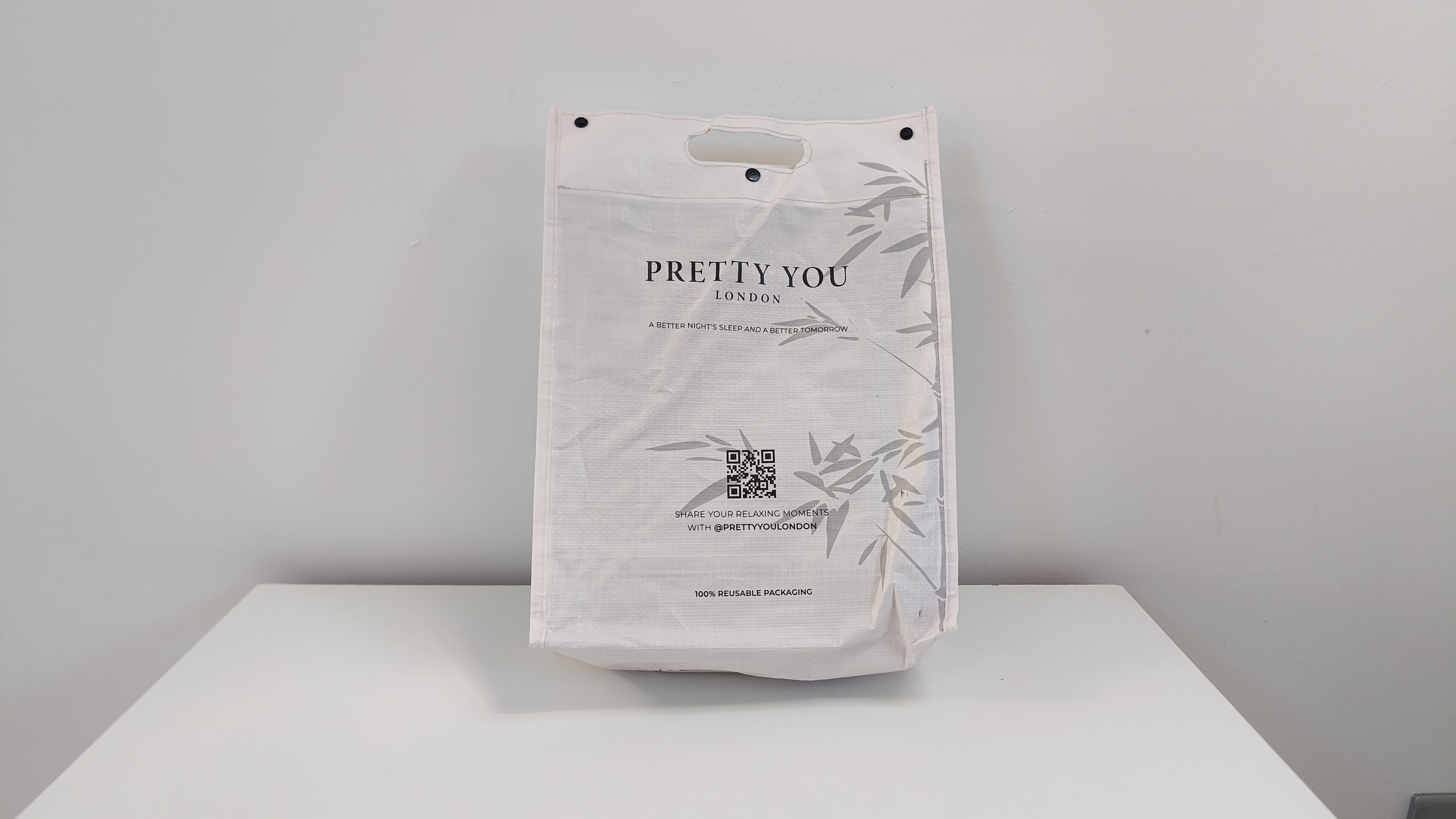 A white, reusable fabric shopping bag with a Pretty You London logo and a subtle grey bamboo leaf print stands upright on a white surface.