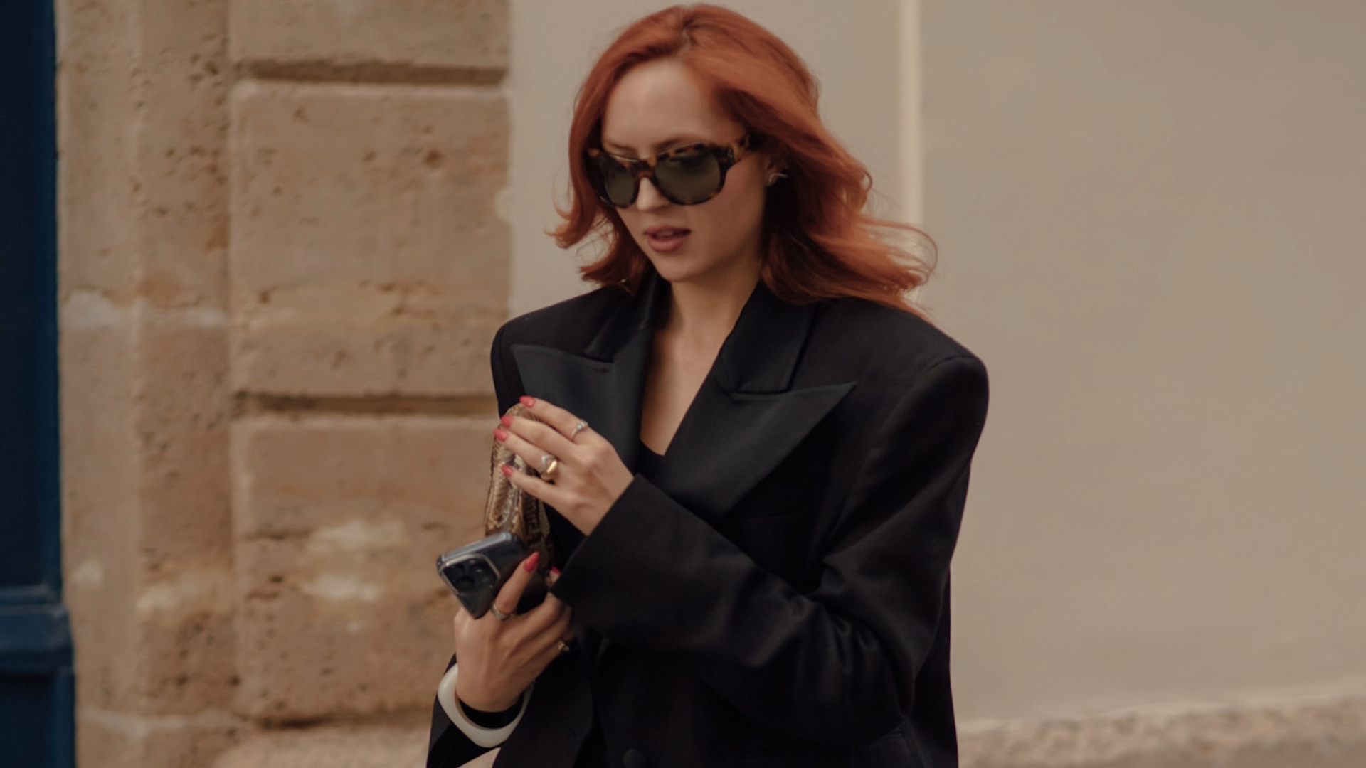 woman with red hair wearing a black blazer and oversized sunglasses. 