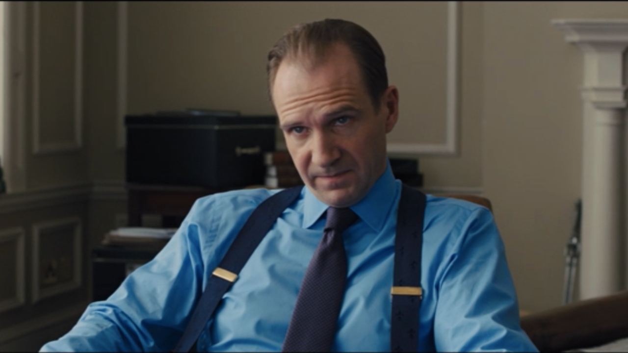 Ralph Fiennes sits down while delivering bad news in Skyfall.