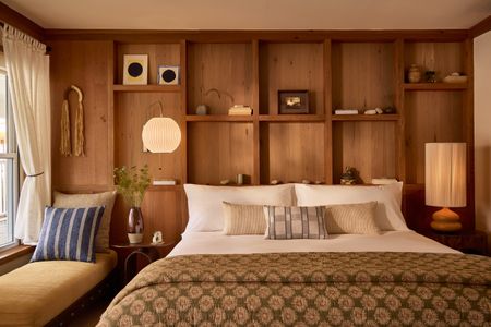 Interiors of a warmly furnished bedroom with ochre, brown, and terracotta tones, a king bed topped with an olive green runner, abstract artwork, warm lighting, and a window seat banquette.