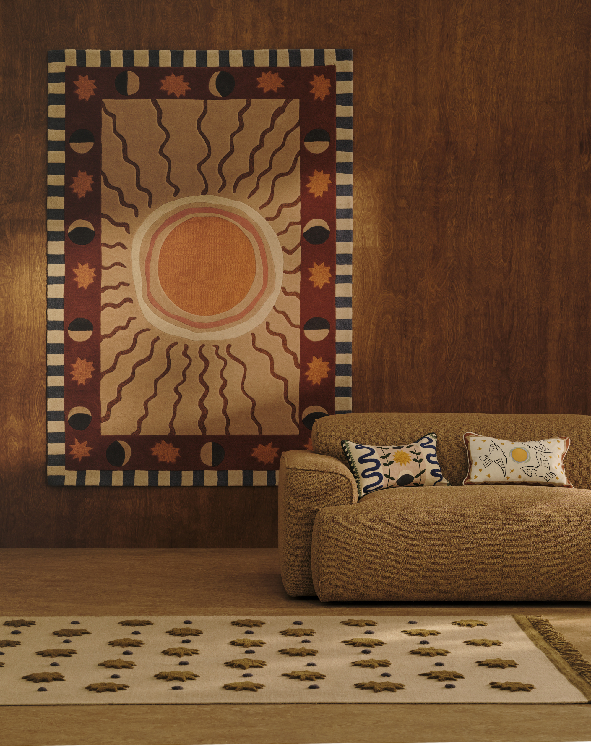 Image of a room with wooden walls and a dark beige floor and matching sofa. There is a colorful rug hung on the wall with a sun on it. And two patterned pillows on the sofa.