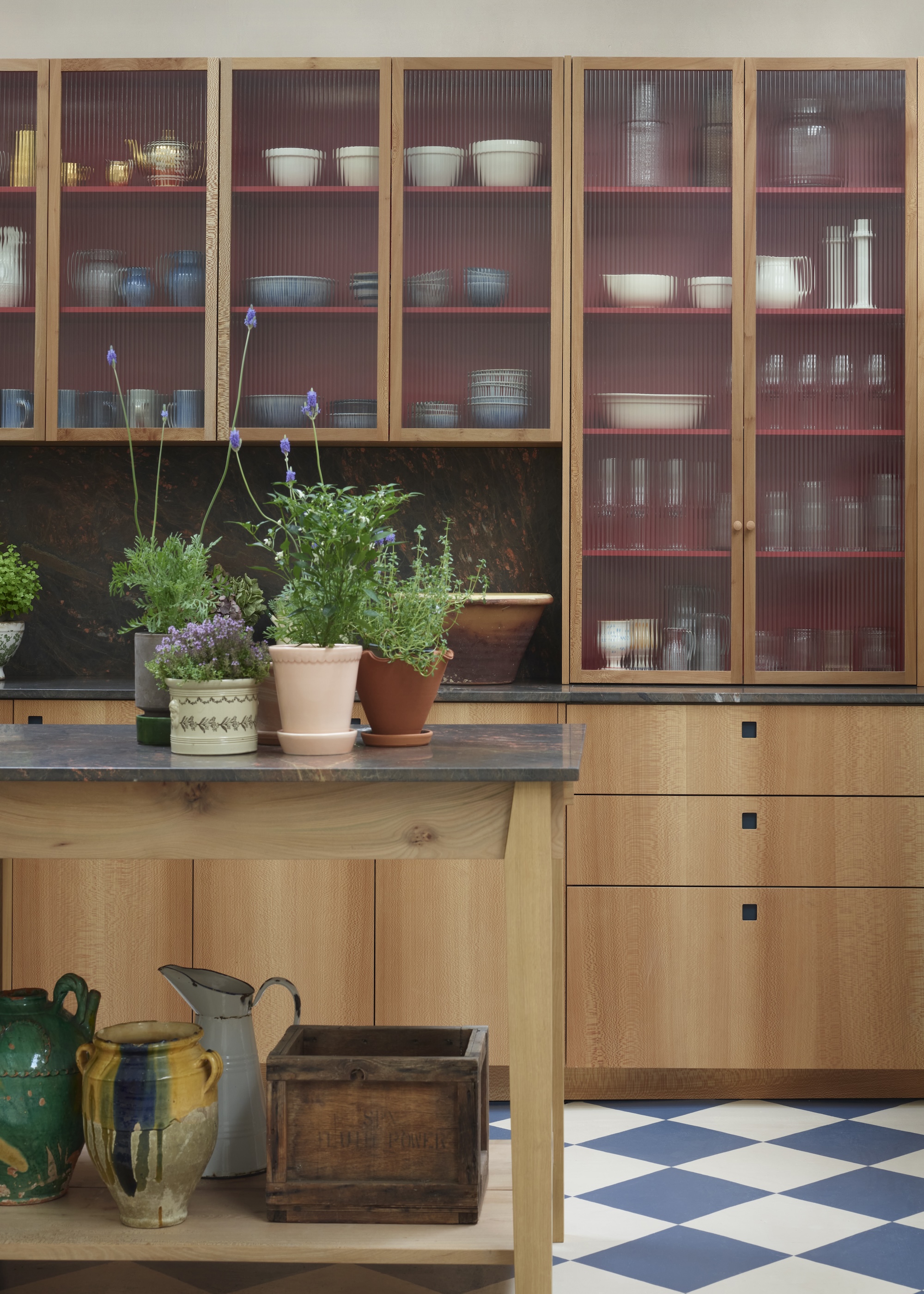 A kitchen with reed glass kitchen cabinet doors with tablware and kitchen essentials inside. There is also a small portable island/table in the middle of the kitchen with plants and pots on top.