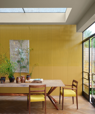 India Yellow painted paneled wall in modern dining room with large wooden table, chairs and rectangular sky light