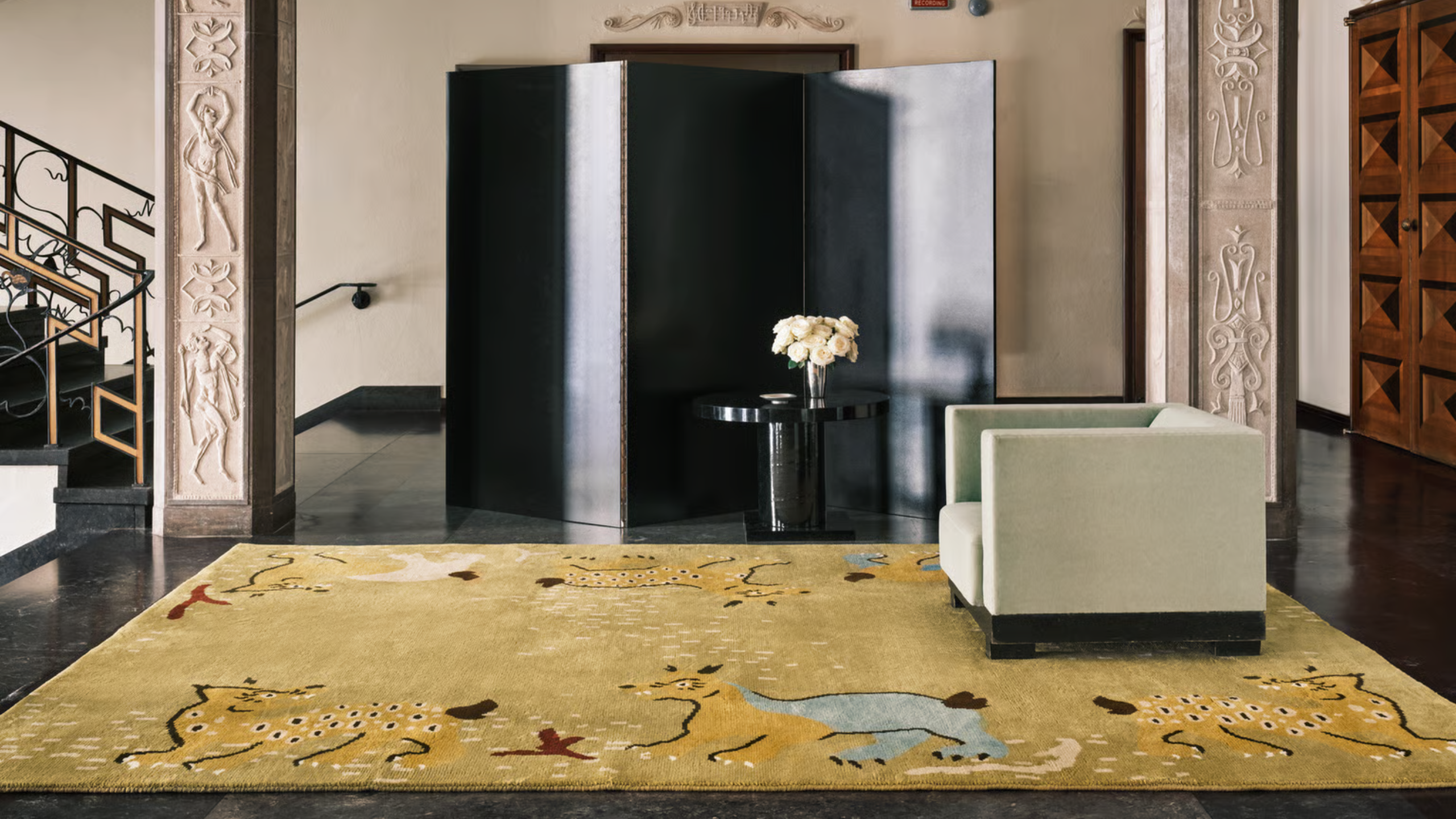 A living room with majestic pillars with moulding, a black glossy room divider, a side table with a vase of flowers, a structured armchair, and a patterned rug with fauna-motifs