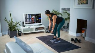 Woman working out with dumbbells in living room, watching TV which is showing a workout on the FIIT app