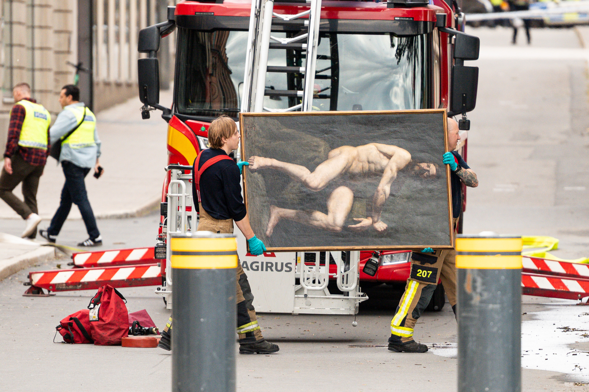 Two firefighters wearing gloves carry a large framed painting of two nude men in front of a fire truck
