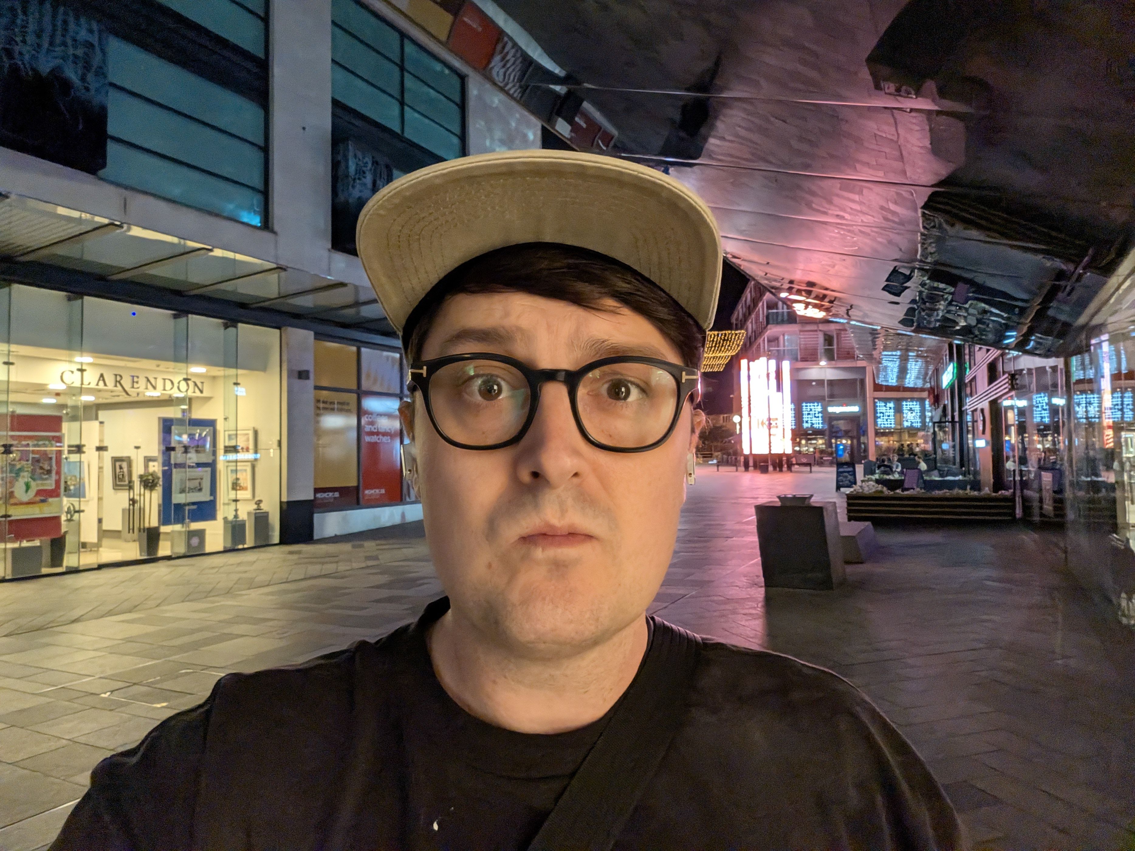 Man wearing glasses and a baseball cap taking a nighttime selfie on a pedestrian shopping street with illuminated signs behind him.