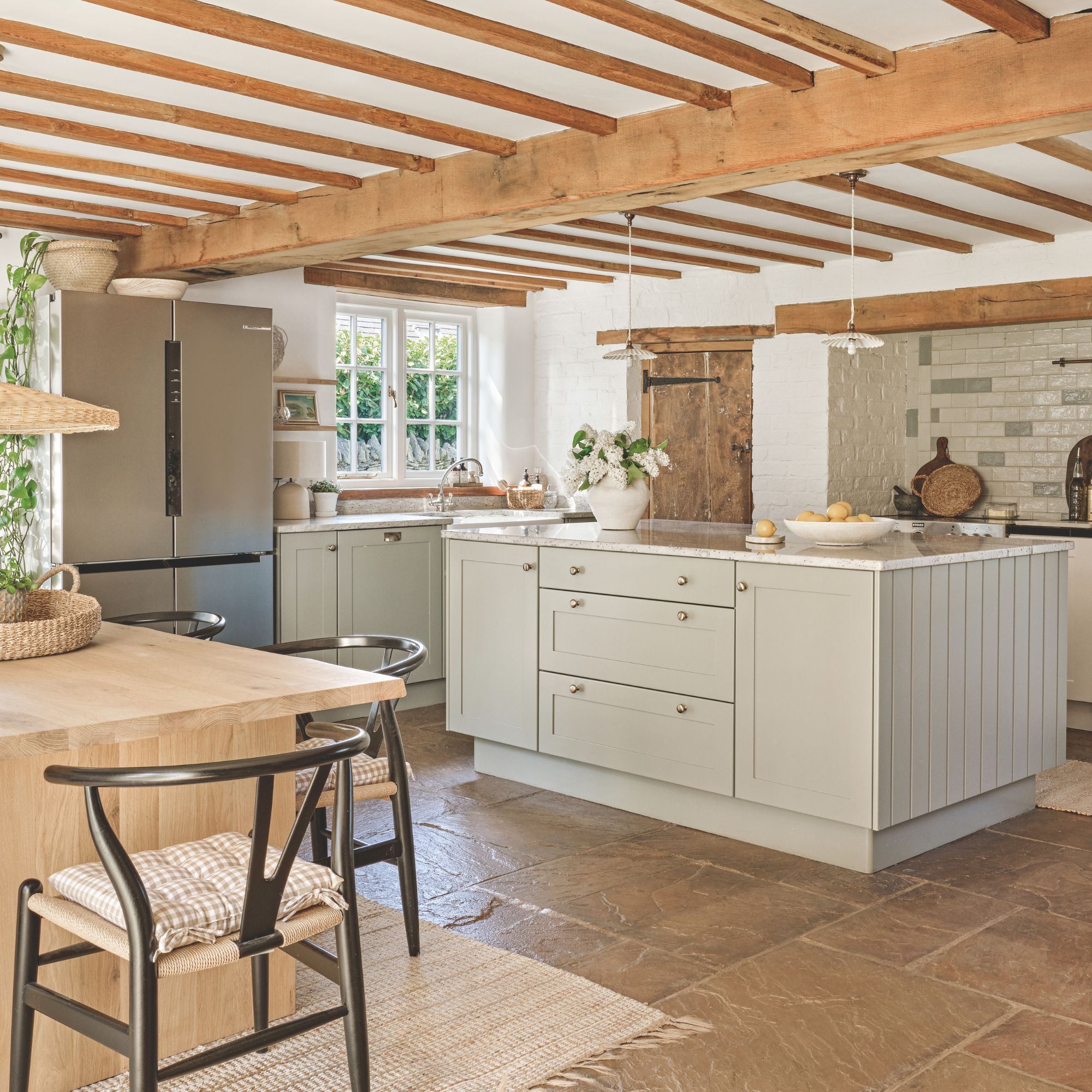 Kitchen diner in cottage with wooden ceiling beams, stone floor and neutral shaker style cabinets. Island with marble top and large American-style fridge freezer. In the corner is a wooden dining table with black wishbone chairs