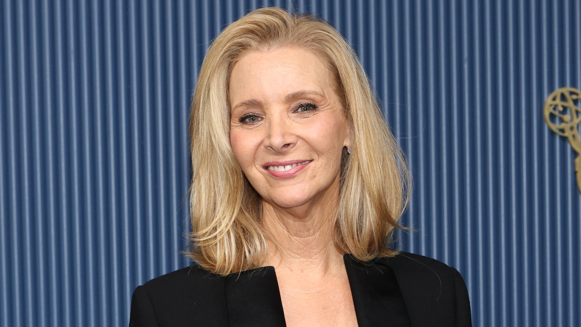 Image of Lisa Kudrow smiling with blonde side-part bob and wearing a black tailored jacket, with blue striped background