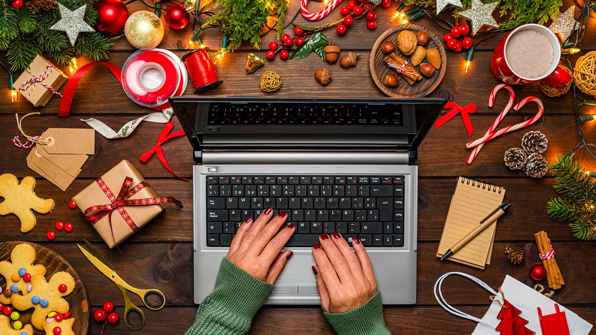 A woman&#039;s arms and hands are shown typing on a laptop on a table. The laptop is surrounded by Christmas decorations, food and gifts