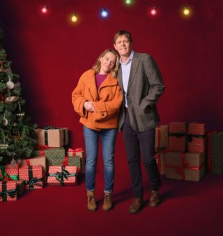 Martha Lloyd (Sally Bretton) and DI Humphrey Goodman (Kris Marshall) stand together in a room with a red backdrop, leaning against eaach other. In the background there is a decorated Christmas tree and several stacks of presents, and a string of multicoloured fairy lights hangs above them.