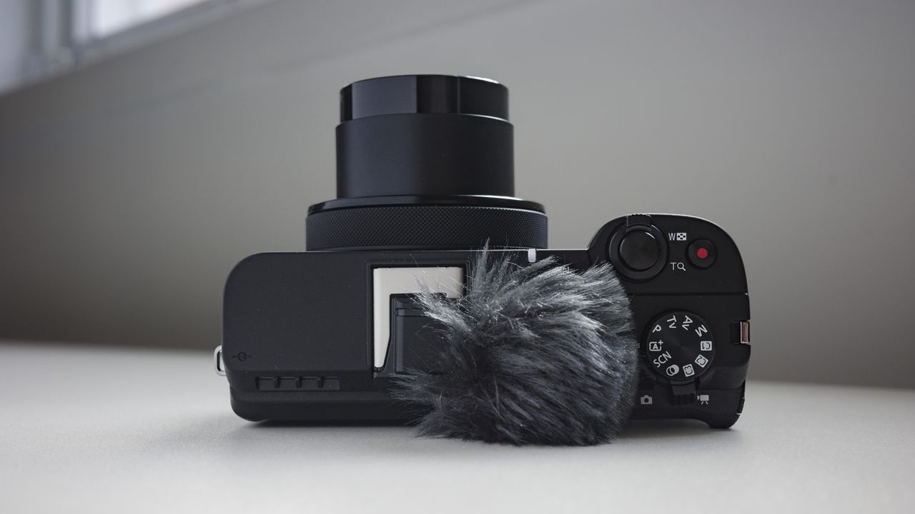 Top plate of the Canon PowerShot V1 compact vlogging camera on a white desk, lens extended, windmuff attached