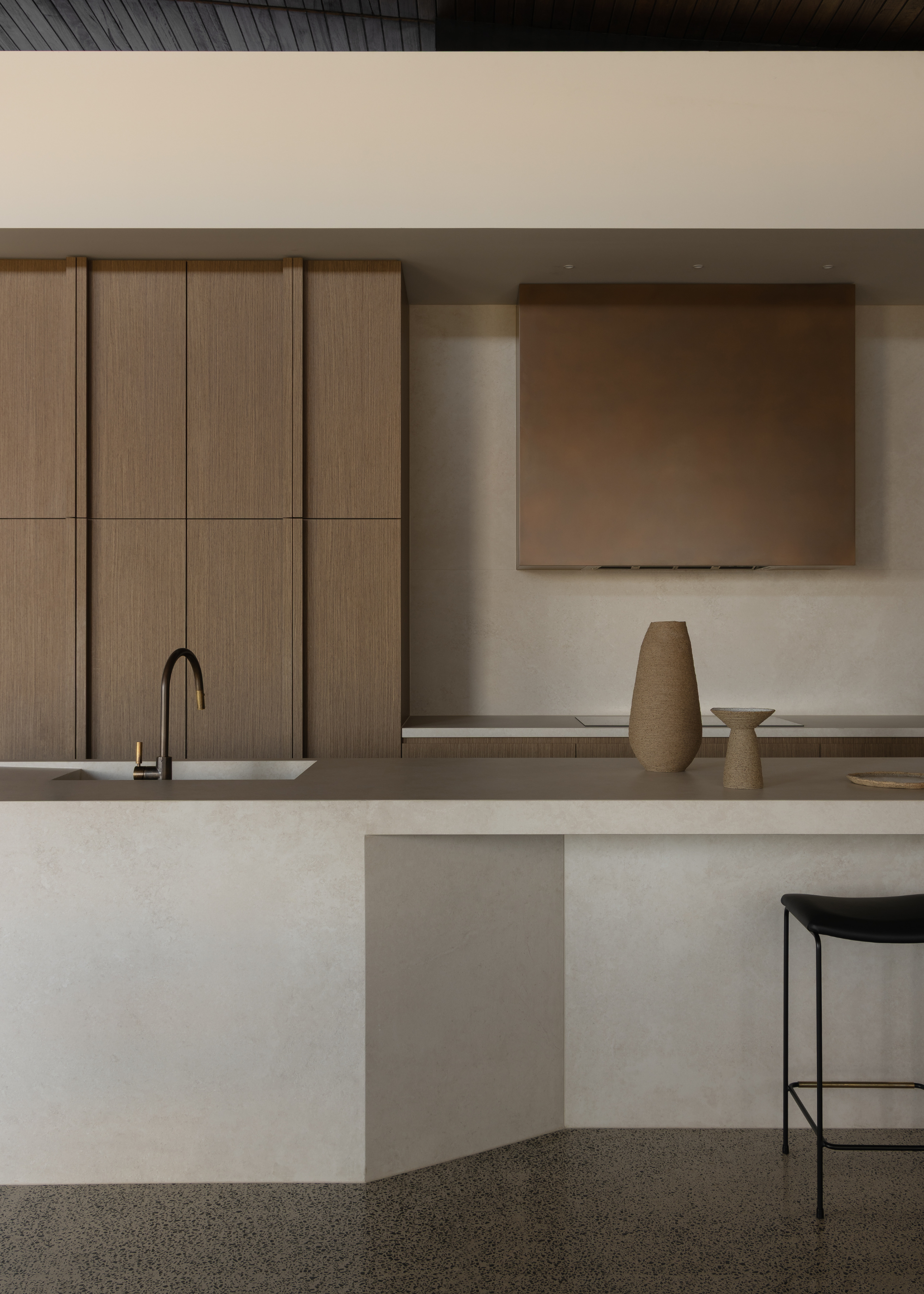 A beige kitchen with wooden cabinets, a stone island, a black stool, and beige ceramics