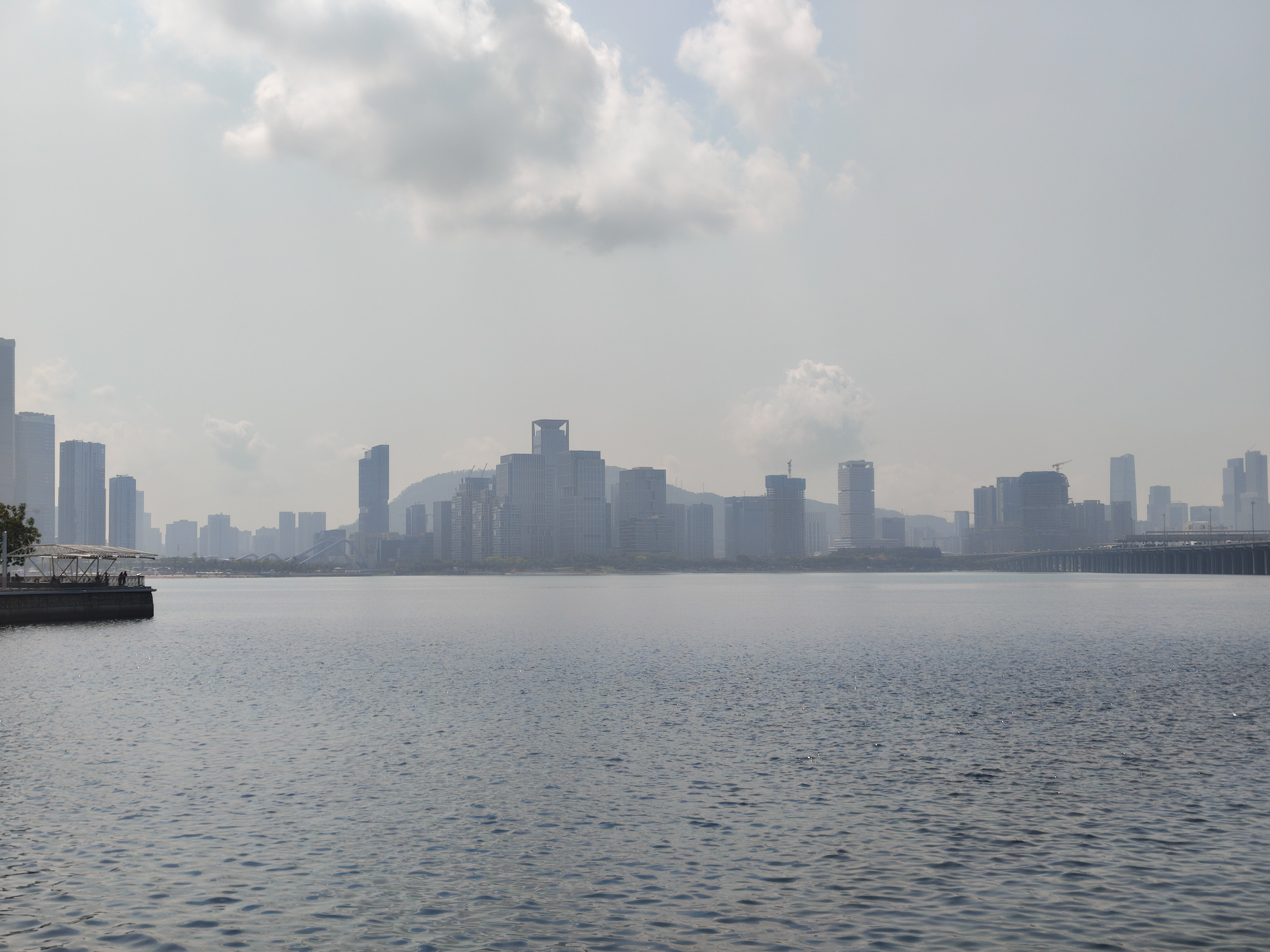 A wide, hazy cityscape across a calm body of water under a bright, cloudy sky. Modern skyscrapers and high-rise buildings line the horizon, with a small pier visible on the far left and a bridge spanning the water on the right.