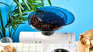 the acaia orion coffee bean doser is a white sci-fi-inspired bean doser with a rotating dial and LED screen and a bean hopper photographed against a blue tom's guide background