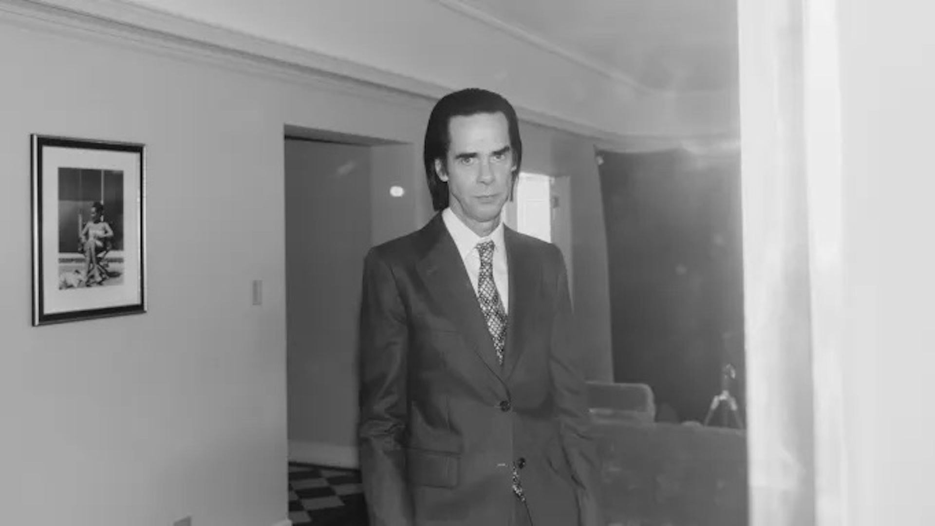 A picture of Nick Cave standing in a suit in a hallway 