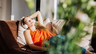 A woman in an orange top napping on sofa next to plants with headphones on listening to binaural beats