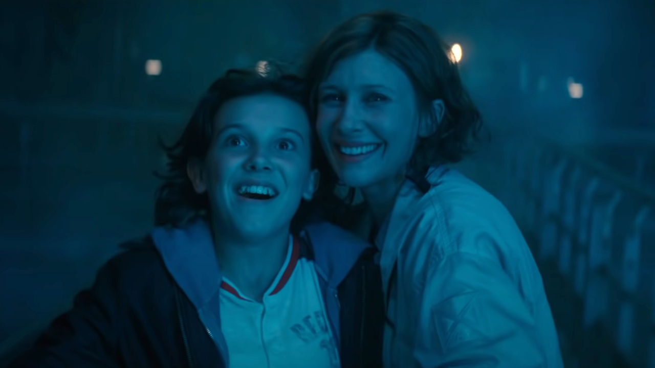 Millie Bobbie Brown and Vera Farmiga stand smiling in a blue light in Godzilla: King of the Monsters.